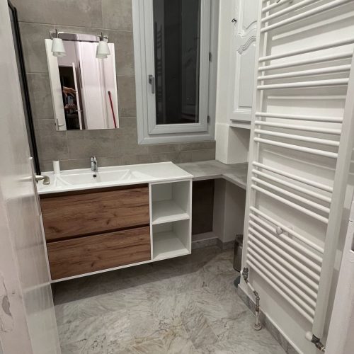 Salle de bain moderne avec meuble suspendu bois, vasque blanche, sèche-serviette, carrelage beige et agencement sur-mesure sous fenêtre