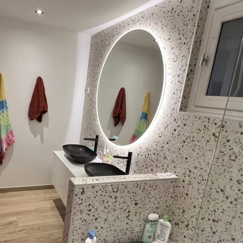 Salle de bain rénovée avec mur en terrazzo, double vasque noire et miroir lumineux rond