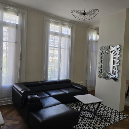Salon rénové dans un appartement haussmannien à Lyon avec parquet en chevron, canapé cuir noir et grandes fenêtres